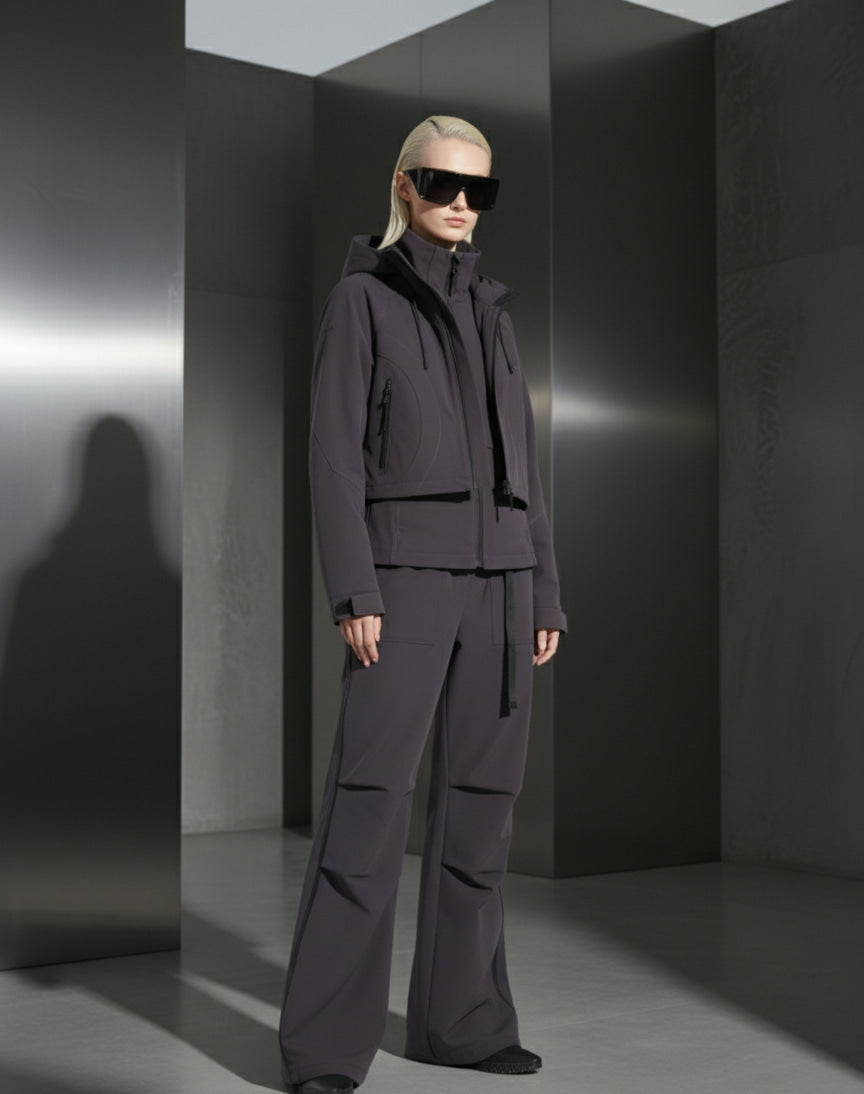 A woman wearing a dark grey outfit and sunglasses against a grey steel background in a minimalistic setting