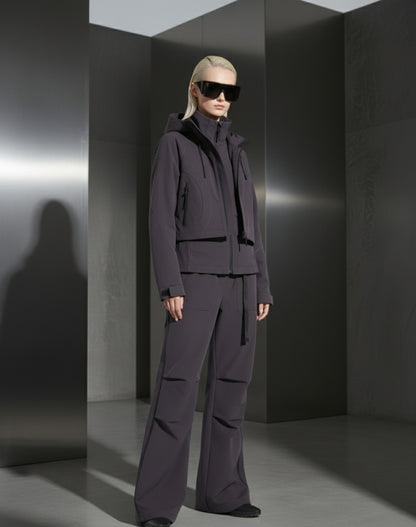 A woman wearing a dark grey outfit and sunglasses against a grey steel background in a minimalistic setting
