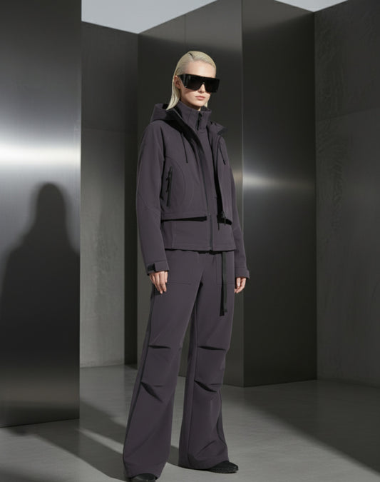 A woman wearing a dark grey outfit and sunglasses against a grey steel background in a minimalistic setting