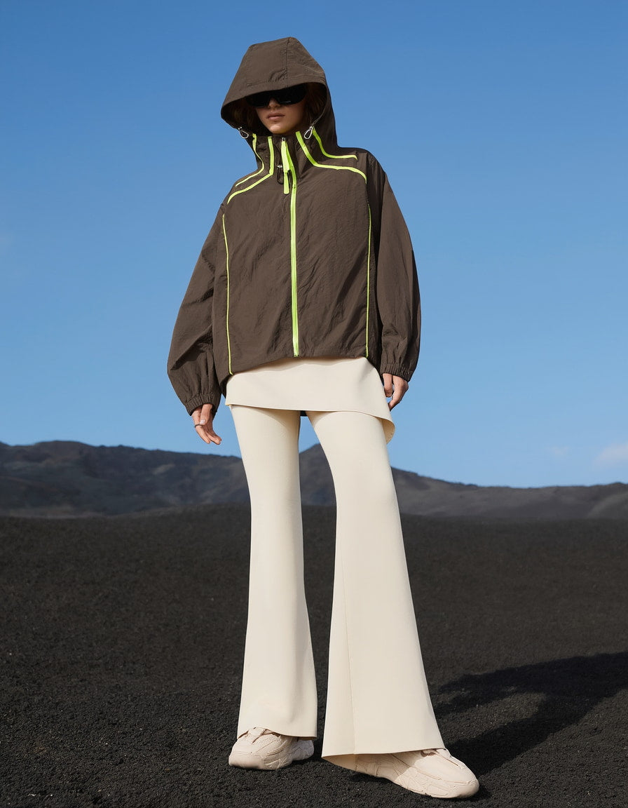 Women wearing a brown jacket activewear with a hood and white pants standing on a black sand beach with mountains in the background.