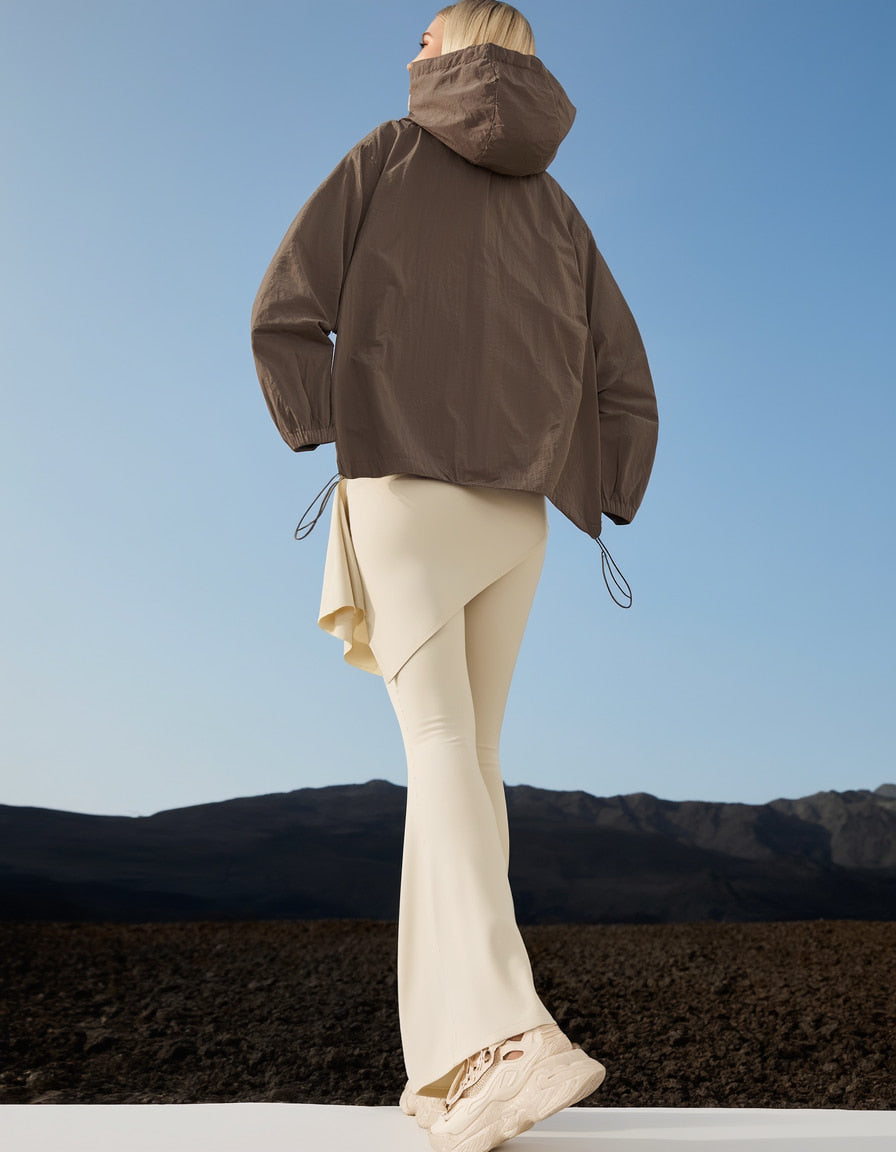 Woman wearing a brown activewear  jacket and beige hot yoga pants standing against a blue sky with mountains in the background